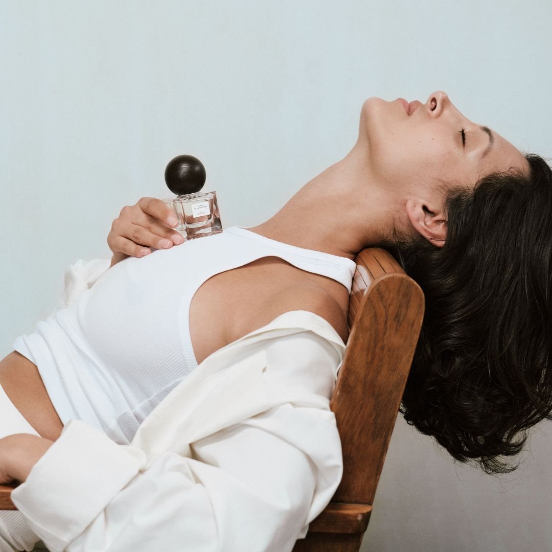 Person holding the 'Main Character' perfume bottle, whilst lying back on a wooden chair with a light background