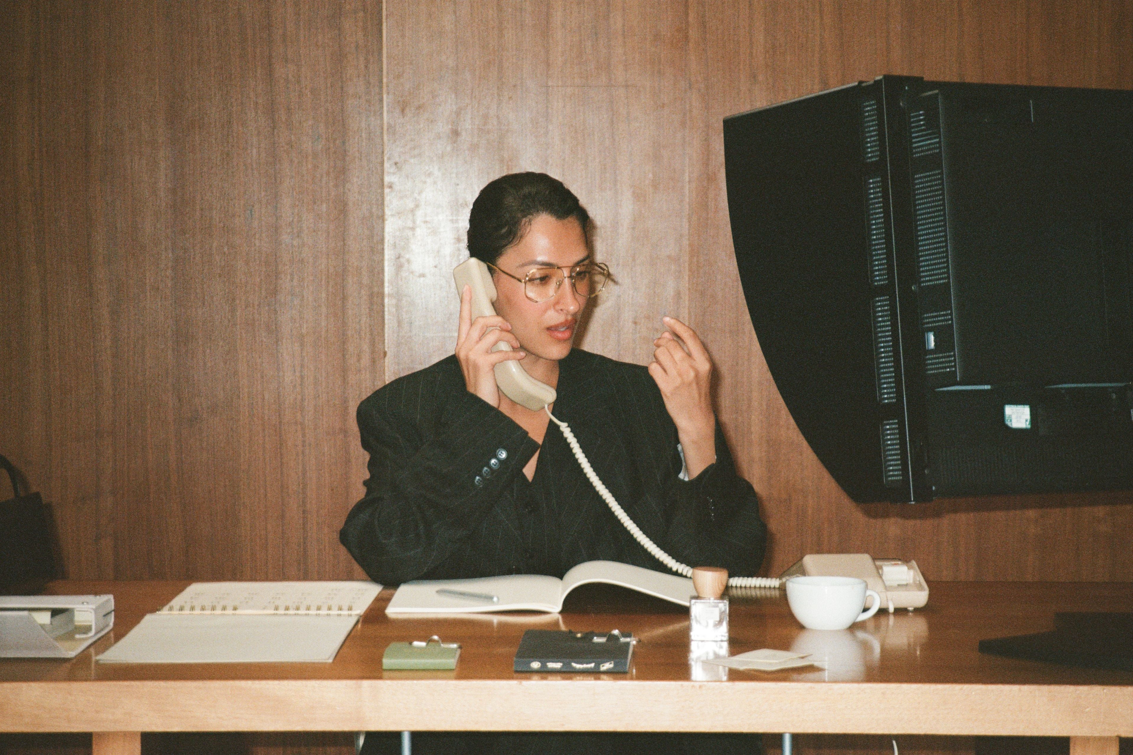 Model sitting at a desk with a computer and phone, wearing glasses.