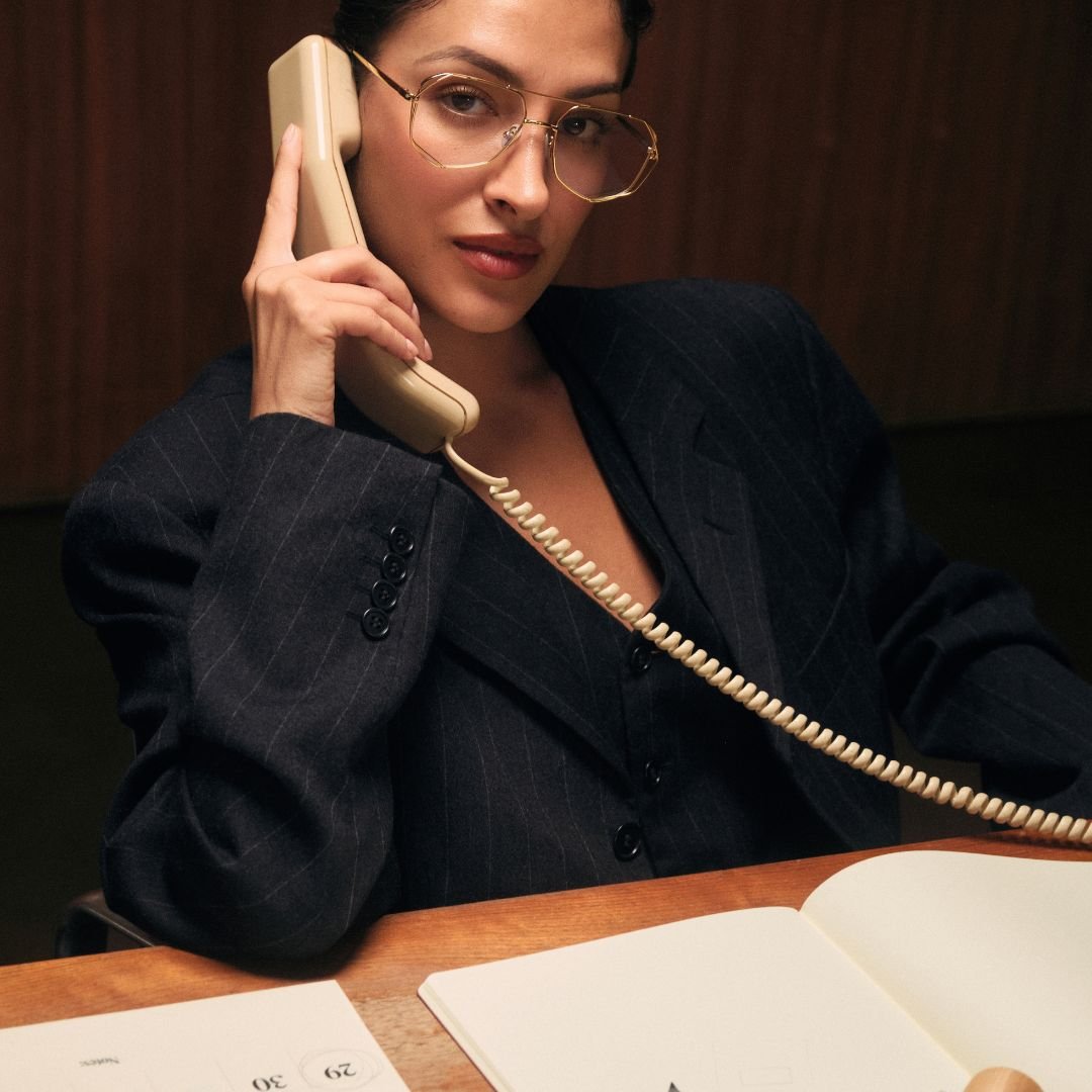 Woman in a black suit talking on a vintage phone whilst sitting at a desk with papers.