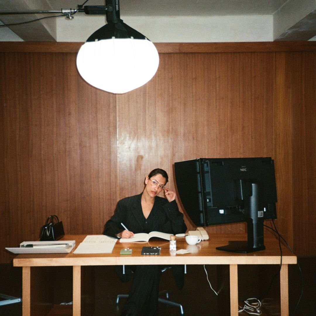 Model sitting at a desk with a computer monitor and various items in a room with wooden walls.