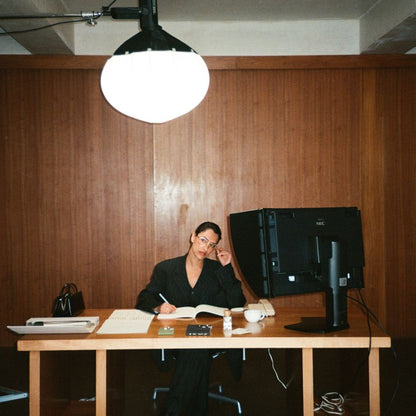 Model sitting at a desk with a computer monitor and various items in a room with wooden walls.