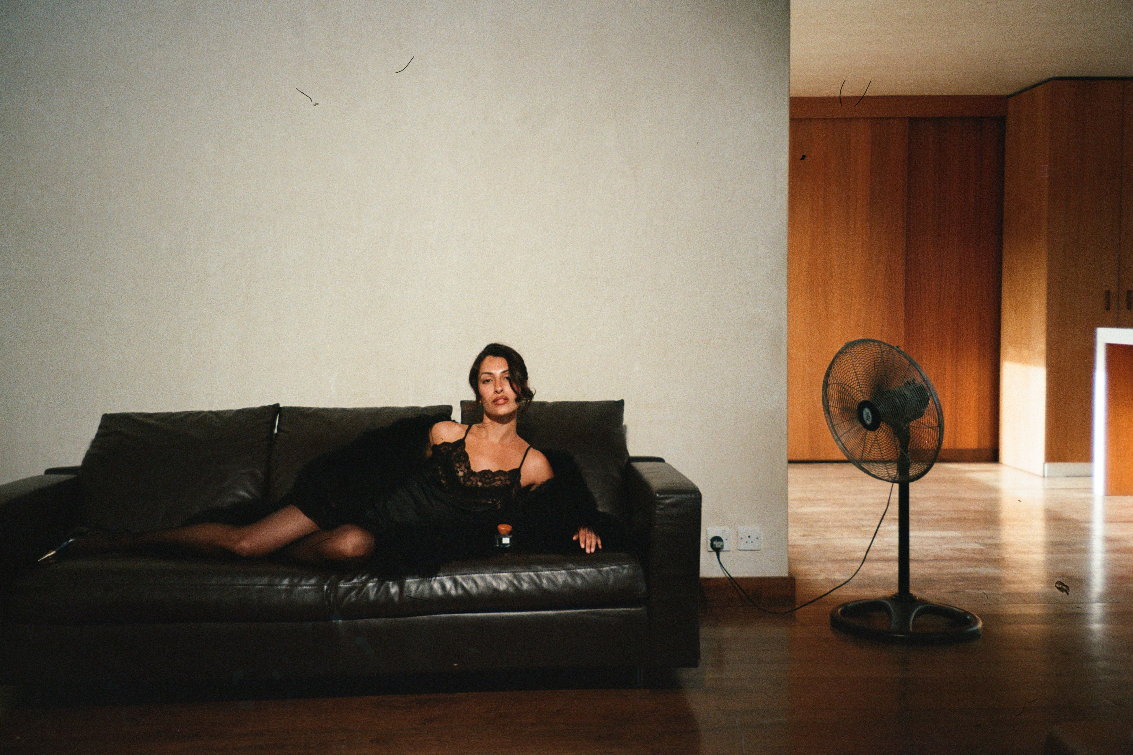 Model laying on a black leather sofa in a room with a fan and wooden floor.
